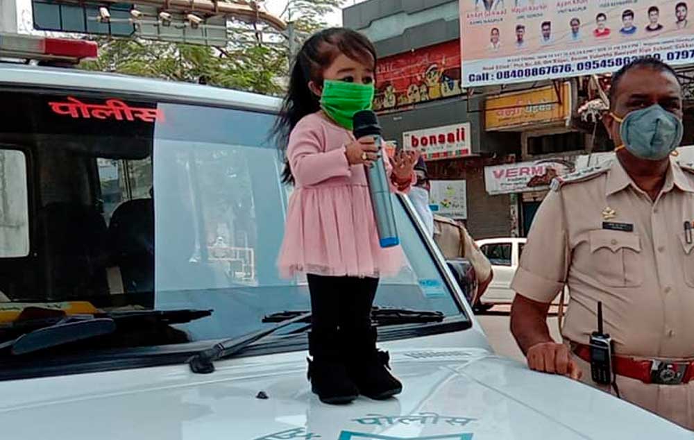 A menor garota do mundo está em cima do capô de um carro de polícia, segurando um microfone, que mede um terço de seu tamanho