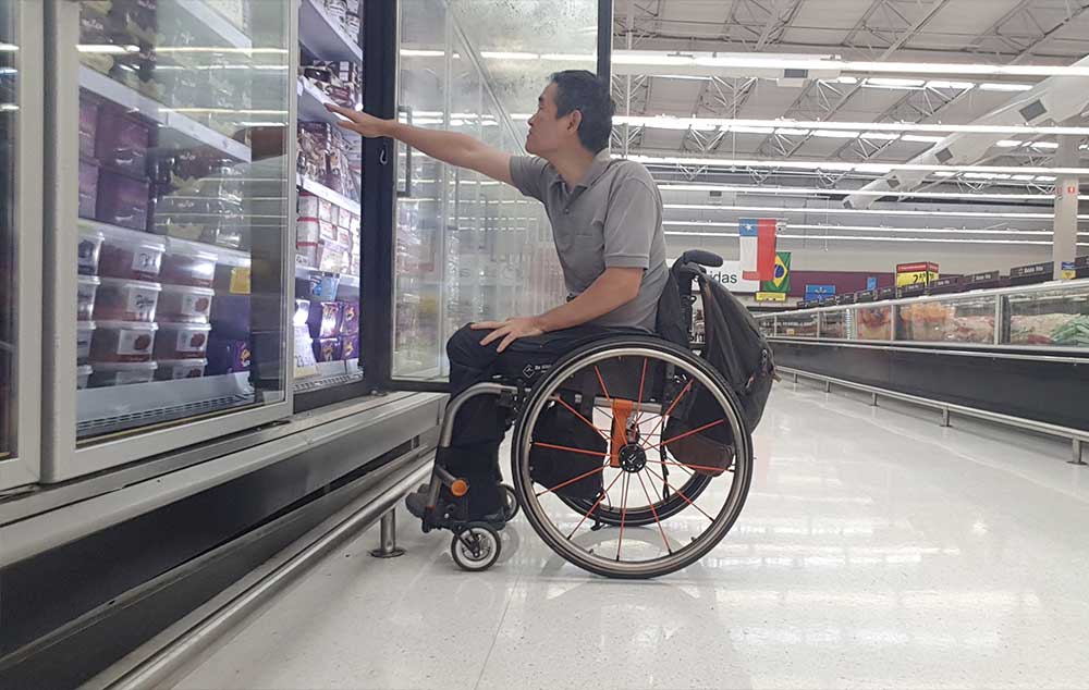 Acessibilidade é fator decisivo. Ricardo Shimosakai está sentado em sua cadeira de rodas, com o braço direito esticado tentando alcançar produtos em uma estante frigorífica de um supermercado