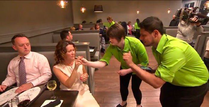 Um casal está sentado em uma mesa de restaurante, sendo atendidos por dois funcionários. A mulher do casal está sendo ensinada pelos funcionários a fazer pedidos em língua de sinais.