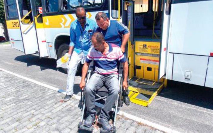 Doi homens auxilian um cadeirante na entrada da plataforma de um ônibus urbano estacionado