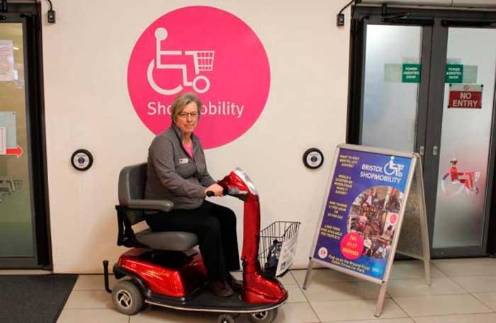 mulher sentada em uma scooter, atrás de um painel escrito "Shopmobility"