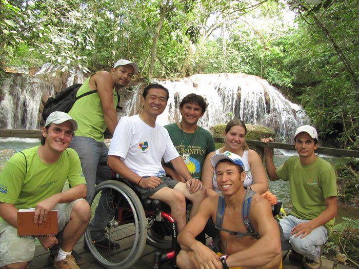 Ricardo Shimosakai está sentado em sua cadeira de rodas, acompanhado de mais 6 pessoas. Ao fundo, uma cachoeira e a mata virgem da cidade de Bonito.