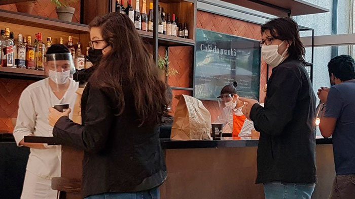 Foto de pessoas em uma fila para pegar sacolas com café da manhã. Estão em um balcão de atendimento do restaurante. Todas estão usando máscara ou face shield.