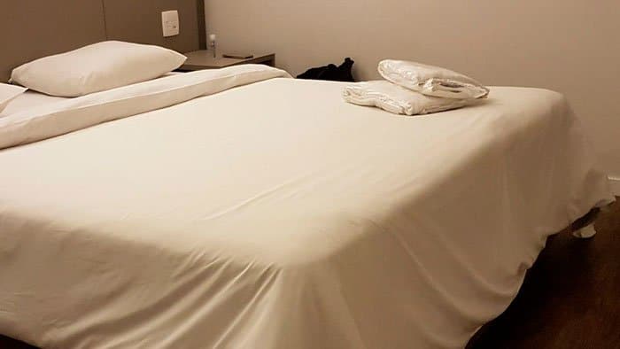 Foto de uma cama de casal com lençol branco. Em cima da cama no canto direito toalhas brancas lacradas em plásticos.