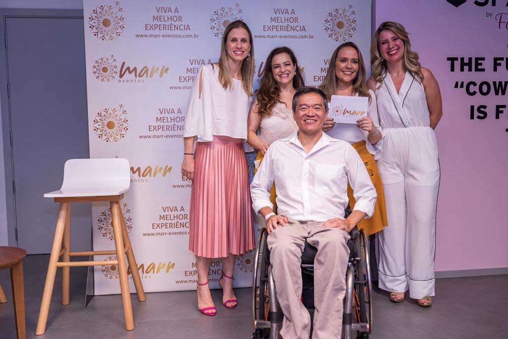 Ricardo Shimosakai está sentado em sua cadeira de rodas, posando para foto com 4 pessoas de pé atrás, na seguinte ordem da direita para a esquerda: Marina Elaine, Karina Pachiega, Maria Ramos e Clarissa Galano. Ao fundo um painel com logos e slogans da Marr Eventos