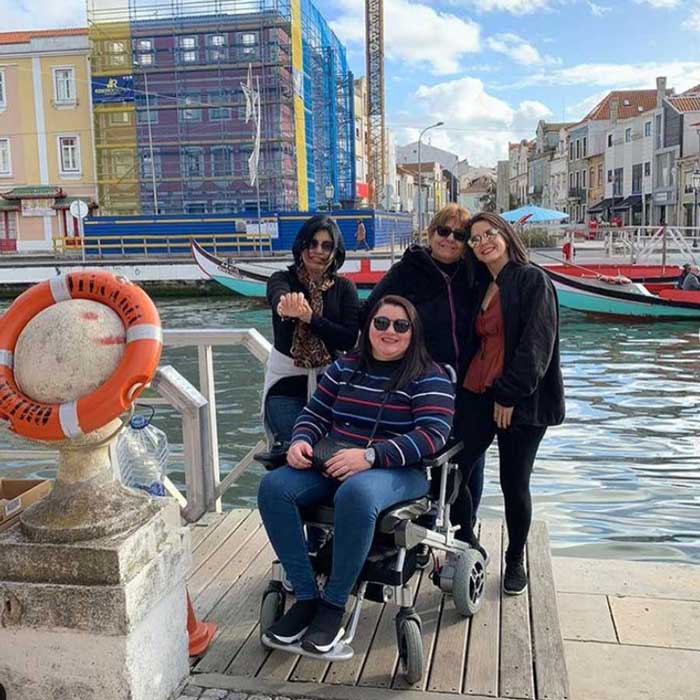 Mulher em uma cadeira motorizada, com três acompanhantes de pé atrás, e ao fundo um canal de água com barcos, margeado por prédios e casas