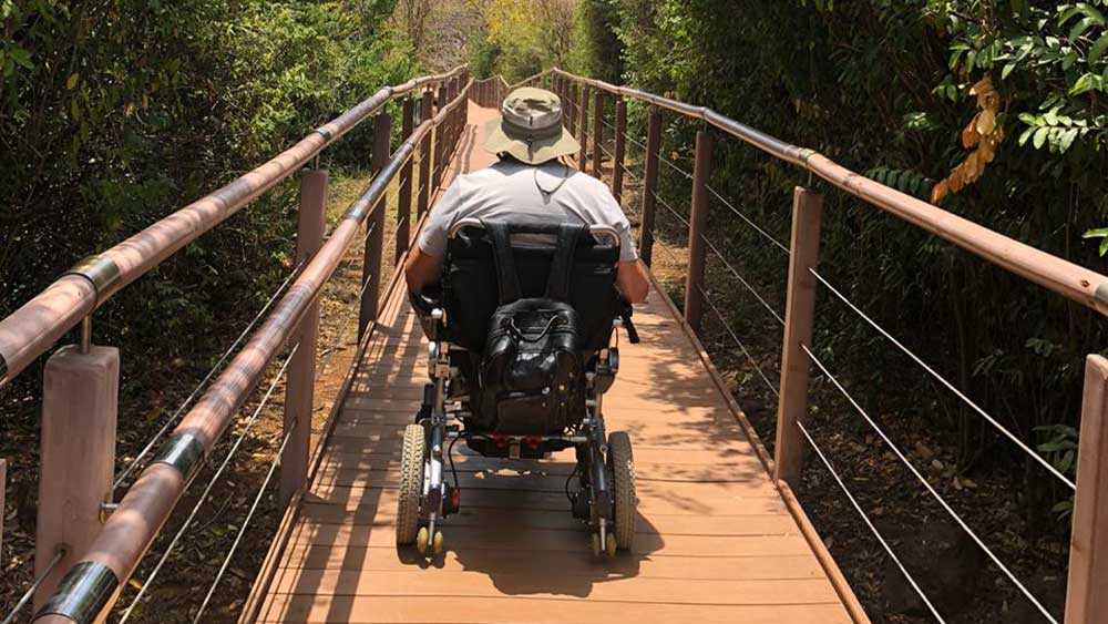Homem em uma cadeira motorizada atravessa uma passarela de madeira com corrimões por dentro de uma local arborizado