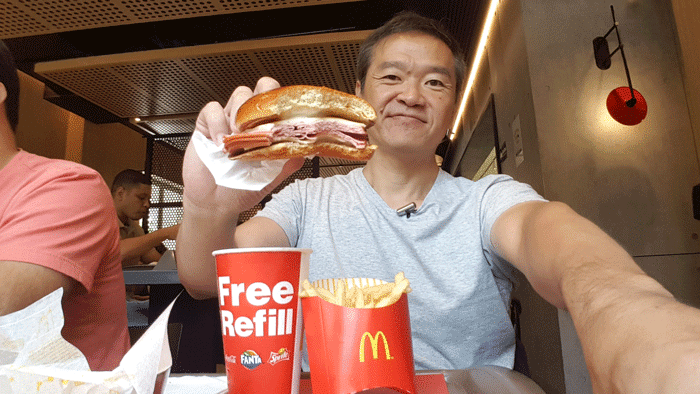 Ricardo Shimosakai segura um hamburguer com a mão direita, e logo na mesa à sua frente, estão um copo de refrigerante e uma caixinha de batatas fritas palito