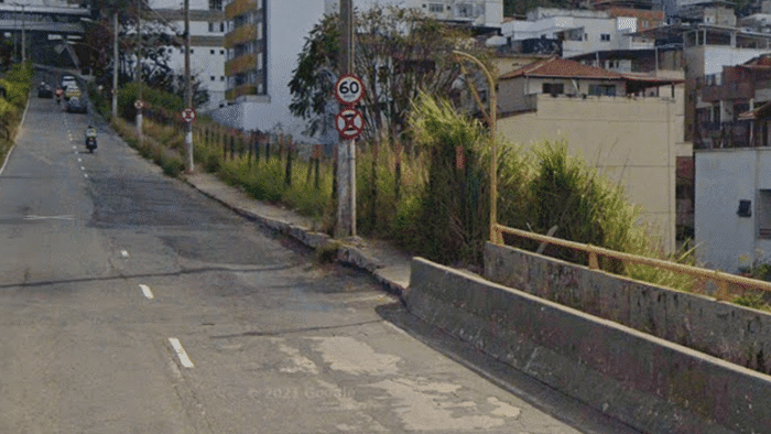 Obstáculos tornam a acessibilidade impossível. Foto mostra o final de uma ponte, com uma calçada estreita, e a visualização da rua extensa e inclinada, sem pontos de travessia de pedestres, enquanto a calçada não tem rebaixamentos de guia, e possui vários postes no meio da passagem.